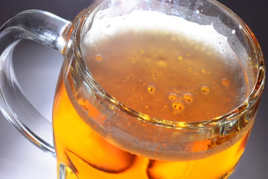 Illuminated mug full of beer with bubbles in light foam on grey background.