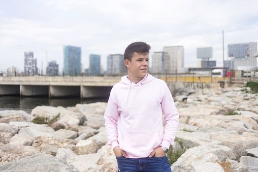 Young teenager male standing against cityscape with hands on pocket on breakwaters while looking away