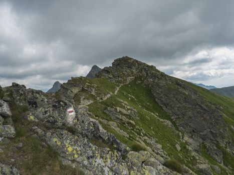 red trail post sign at steep hiking path on ridge of rohace mountain to peak ostry rohac. Sharp green rocks, .Summer blue sky white clouds, Slovakia, Western Tatra mountain