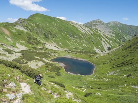 Men hiker walking down to B.eautiful vivid blue mountain lake Horne Jamnicke pleso with green mountain peaks view from the Jamnice sedlo saddle, Western Tatras mountains, Rohace Slovakia, summer sunny day blue sky