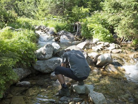 Young men hiker refreshing his head at mountain stream creek flows between lush green fern leaves and boulders. Western Tatras mountains, Rohace Slovakia, summer.