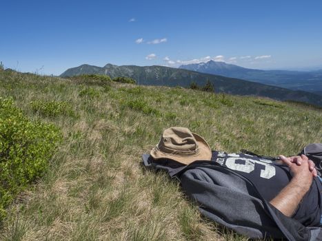man hiker in hat taking a nap at meadow with Mountain landscape of Western Tatra mountains or Rohace with view on high tatras with Krivan peak from hiking trail on Baranec. Summer blue sky background.