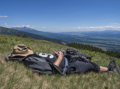 man hiker in hat taking a nap at meadow with Mountain landscape of Western Tatra mountains or Rohace with view on high tatras with Krivan peak from hiking trail on Baranec. Summer blue sky background.