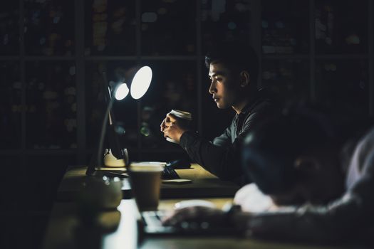 Asian businessman working hard late with his technology laptop in office, customer service and call center department, hard worker and overtime, her colleagues sleepy, low light environment,