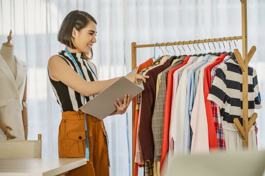 Young Asian designer woman choosing and checking multicolor clothes with checklist at workplace,small business startup, Business owner entrepreneur, modern freelance job lifestyle concept. asia people