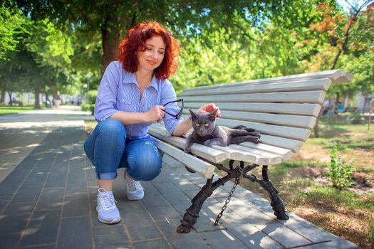 A red-haired curly girl in the park on a bench stroking a gray street cat. Love to the animals