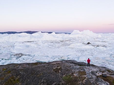 Travel in arctic landscape nature with icebergs - Greenland tourist explorer. Person looking at amazing Greenland icefjord - aerial photp. Man by ice and iceberg in Ilulissat. Climate change concept.