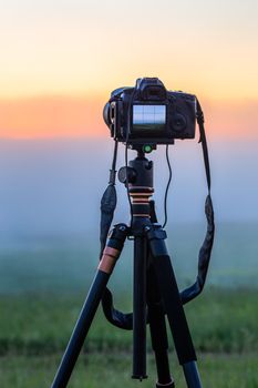 black digital camera on tripod shooting foggy morning landscape at summer with selective focus.