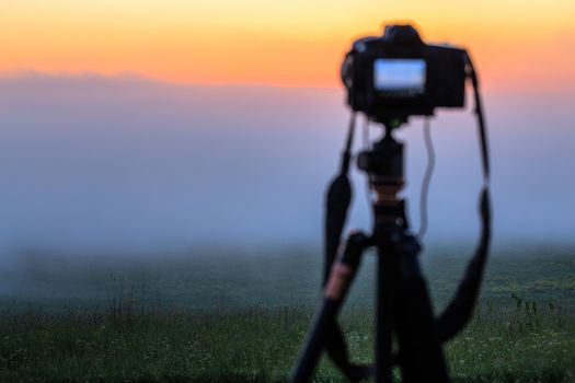 black digital camera on tripod shooting foggy morning landscape at summer with selective focus.
