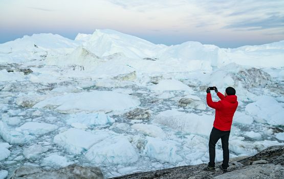 Travel in arctic landscape nature with icebergs. Greenland tourist man explorer taking photo with phone of amazing Greenland icefjord in Ilulissat affected by climate change and global warming.