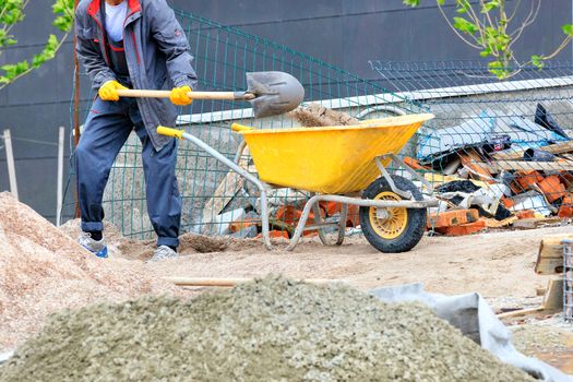 A worker in a dark blue overalls with a shovel loads sand in a yellow construction wheelbarrow against the background of a working construction site.