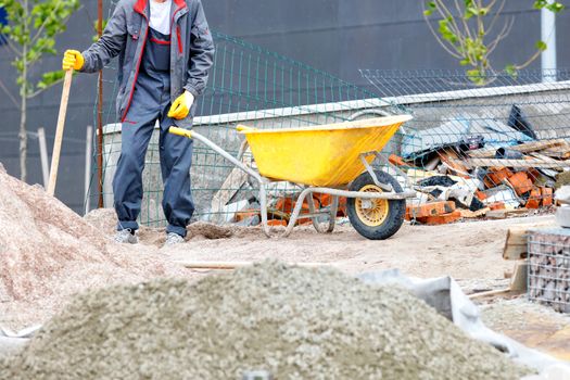 A worker in blue overalls stands near a pile of sand and a yellow construction wheelbarrow against the background of a construction workplace, image with copy space.