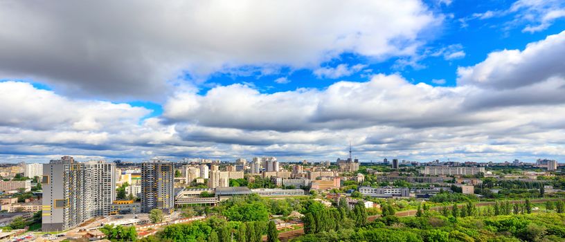 Large low clouds fill the sky over a sunny city with tall apartment buildings and green parks in a panorama with cityscape and horizon.