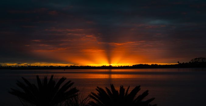 Brilliant sunrise through cloudy sky with rays of orange light reflecting on water in bay.