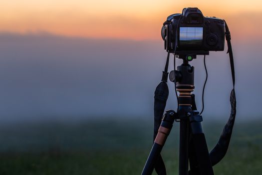 black digital camera on tripod shooting foggy morning landscape at summer with selective focus.