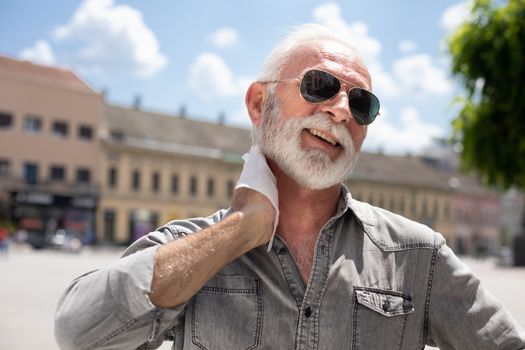 Old man cleaning neck and sweat with wet wipes on street on hot summer day, blurred urban city background