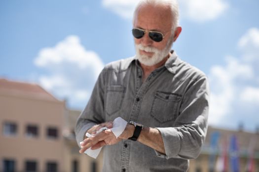 Old man cleaning hands with wet wipes outdoor in public, hot summer day, sunglasses