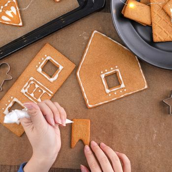 Young woman is decorating Christmas Gingerbread House cookies biscuit at home with frosting topping in icing bag, close up, lifestyle.