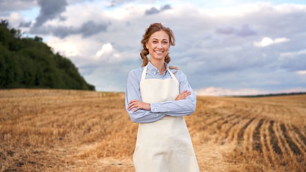 Woman farmer apron standing farmland smiling Female agronomist specialist farming agribusiness Happy positive caucasian worker agricultural field Pretty girl arms crossed cloudy sky background
