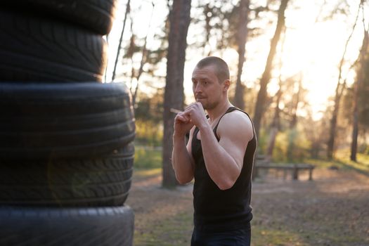 Man fighter training boxing outdoor tire DIY handmade gym in forest fitness workout. Young adult training right hand hit nature without boxing gloves. Sportive activie male healthy lifestyle concept.