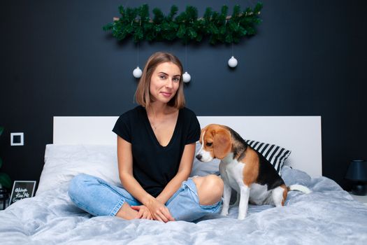 Young beautiful woman playing with dog sitting on bed in a stylish bedroom. Domestic animals at home. Beagle dog. Friendship. Comfort cosiness