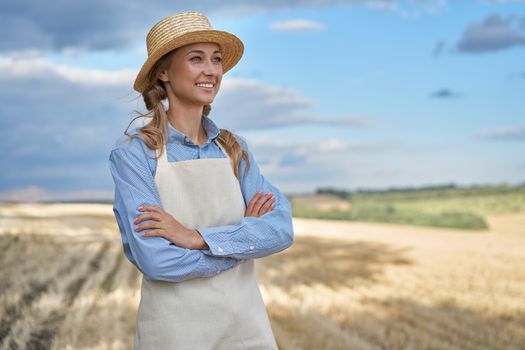 Woman farmer straw hat apron standing farmland smiling Female agronomist specialist farming agribusiness Happy positive caucasian worker agricultural field Girl arms crossed cloudy sky background