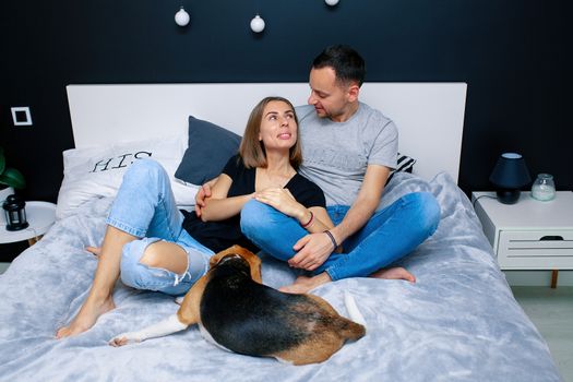 Young couple sitting on a bed in the bedroom, hugging. Near them is their dog. Domestic animal Beagle