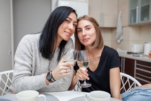 Friends meeting with wine and cake in the modern style kitchen. Young women smile and joke with glasses of wine in his hands. Two girls drink wine in the home kitchen.
