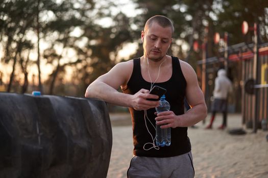 Young adult caucasian athlete listening phone music white headphones after workout. Handsome sportsman resting after cross training exercises sunset background. Healthy lifestyle concept. Drink water