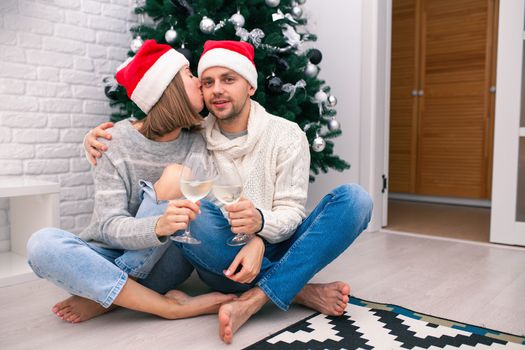Young happy couple in Christmas hats near a Christmas tree kissing, holding glasses of wine. New Year celebration. Happy New Year Family Concept. Christmas for two