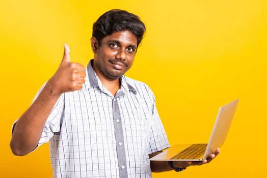 Asian happy portrait young black man smiling standing wear shirt using laptop computer and showing thumb up looking to camera isolated, studio shot yellow background with copy space