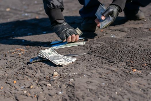 view of money and a knife lying on the asphalt against the background of the hands of a bandit who picks up bills from the road