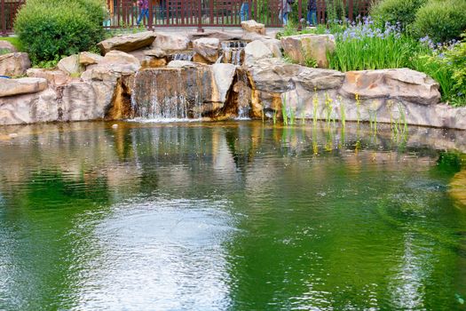 Reflection of the sky and greenery in the pond of a city park, a decorative waterfall pours between large stone boulders, copy space.