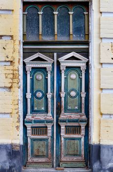 Old tall wooden entrance doors in blue and white colors with carved elements are framed on the facade of an old house with a carved symmetrical pattern.