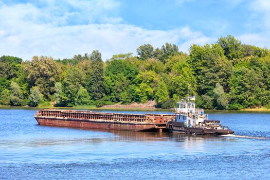 River tugboat pushes an empty rusty barge along the river against the background of coastal greenery and calm surface of the water, the concept of river freight transportation, copy space.