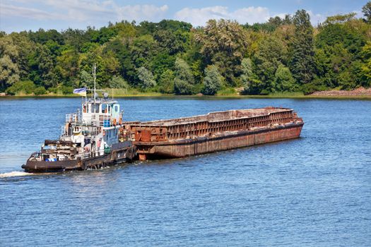 River tugboat pushes an empty rusty barge across a wide river to the other bank against the backdrop of coastal greenery and a surface of the water, the concept of river freight transport, copy space.