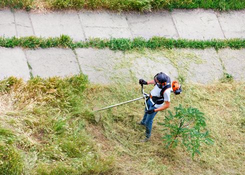 A service worker with a petrol industrial trimmer mows tall grass on the lawn of a city park along a pedestrian walkway. Copy space, top view.