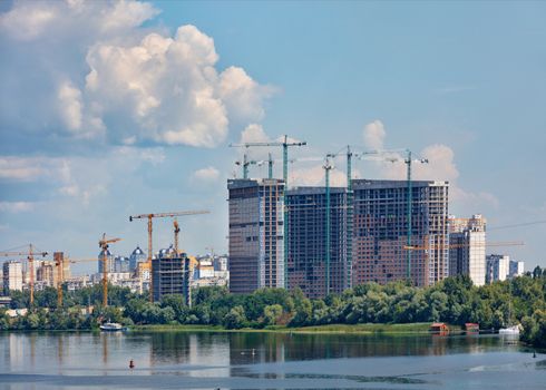 Construction site on the river bank, facades of high-rise residential buildings under construction with tower cranes against the background of a blue cloudy sky, copy space.