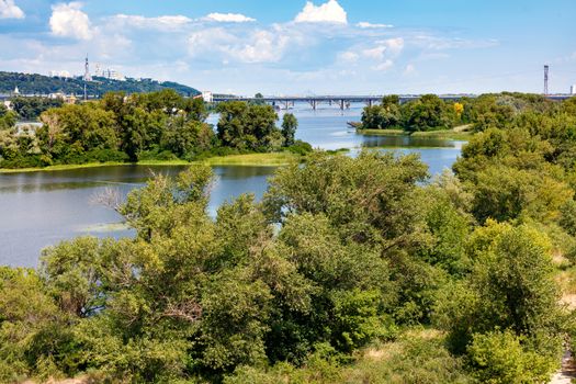 The picturesque islands on the Dnipro River are overgrown with trees and green grass. View of the bridges over the river and the hills of Kyiv.