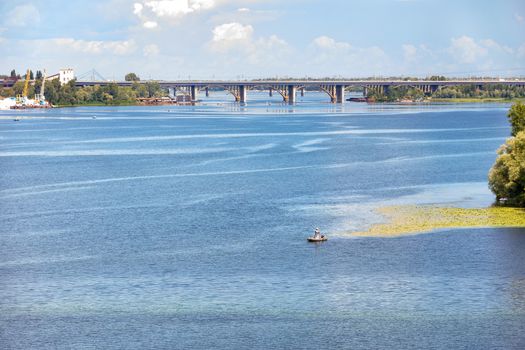 The water surface of the wide Dnipro River in the foreground, a lone fisherman on an inflatable boat in the middle, road bridges across the river on the horizon, copy space.
