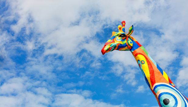 Painted cheerful giraffe head against a blue, slightly cloudy sky in sunlight, copy space.