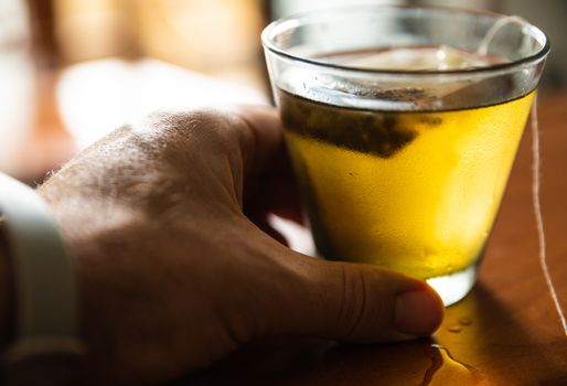 Close-up image of a cold herbal tea: the glass is covered with condensation. The sachet is infused in the glass. A Caucasian man's hand grabs the glass