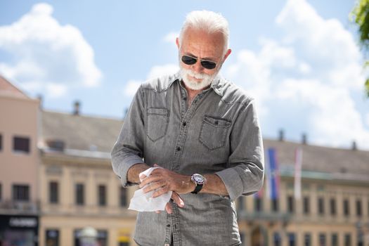 Old man cleaning hands with wet wipes outdoor in public, hot summer day, sunglasses