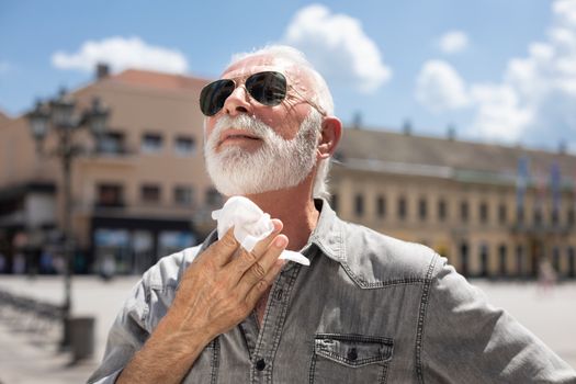 Old man cleaning neck and sweat with wet wipes on street on hot summer day, blurred urban city background
