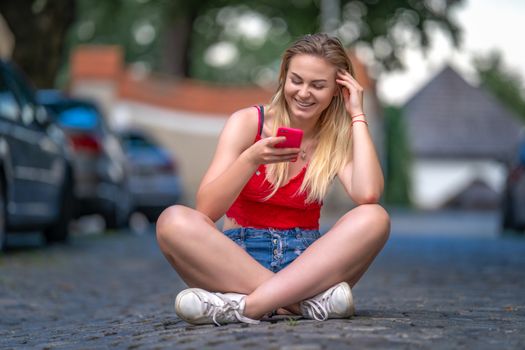 portrait of a beautiful young woman sitting on the pavement in the city streets using a smartphone.