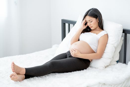 Beautiful Asian pregnant woman show action of stress and headache lie on bed with morning light. Concept of sickness of mother during pregnancy and need to take care and support at home.