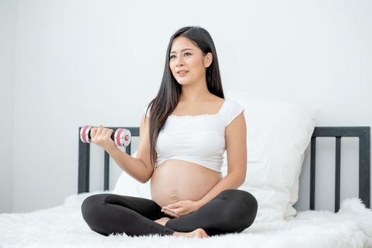 Portrait of Asian pregnant woman hold dumbbell and smile also sit on bed with morning light. Concept of good healthcare support baby in the womb and stay safe at home.