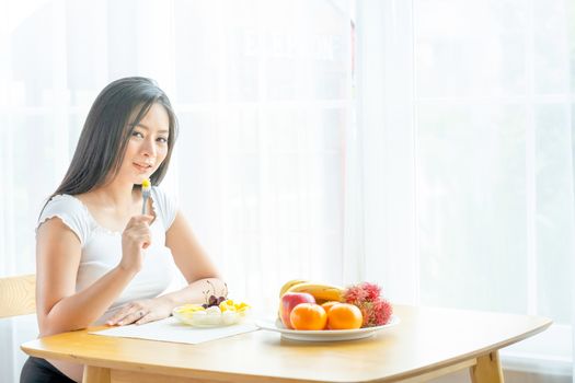 Beautiful pregnant woman hold folk with some fruits and sit near table with various types of fruits in front of curtain with morning light. Concept of good and healthy food for pregnant people.