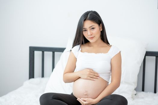 Portrait of beautiful Asian pregnant woman sit on bed and touch and look to her belly also smiling. Concept of good take care baby in the womb of healthy mother and stay at home.