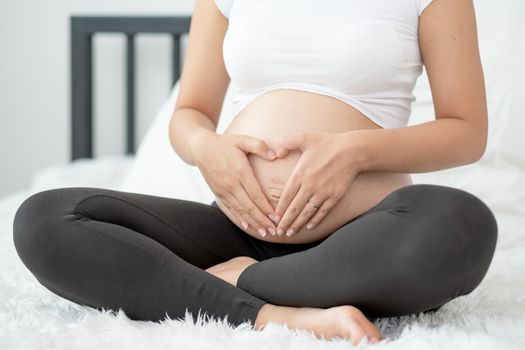 Close up pregnant woman hands touch her belly and make symbol of heart also sit on white bed with morning light. Concept of good take care baby in the womb of healthy mother and stay at home.